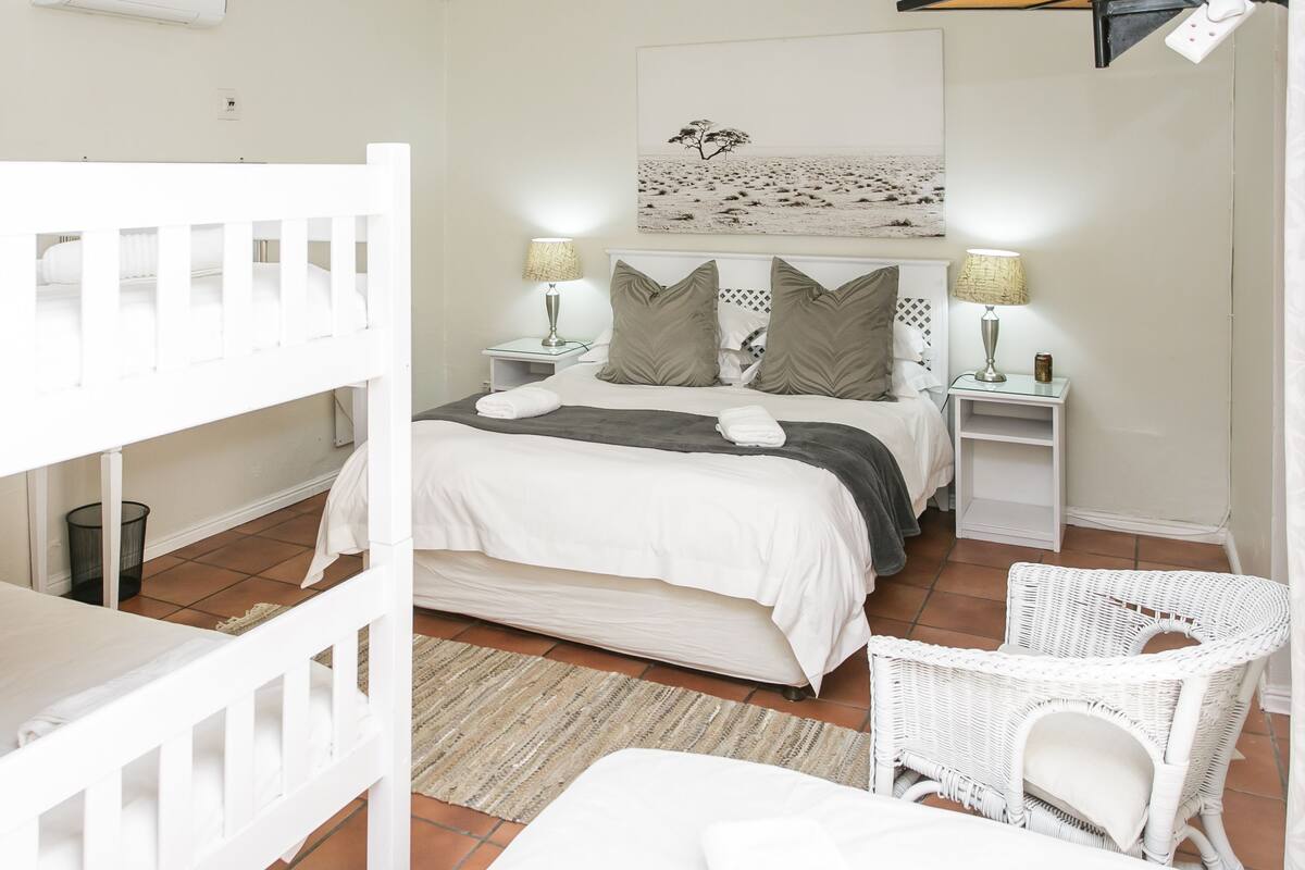 A bedroom with a bed, two nightstands, a white chair, a crib, and a painting on the wall.
