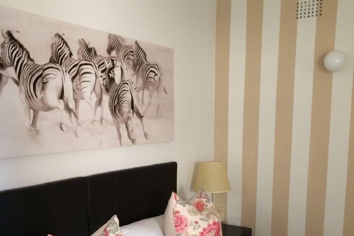 A hotel room with a striped wall, a zebra painting, a bed with floral pillows, a nightstand with a lamp, and a towel on the floor.