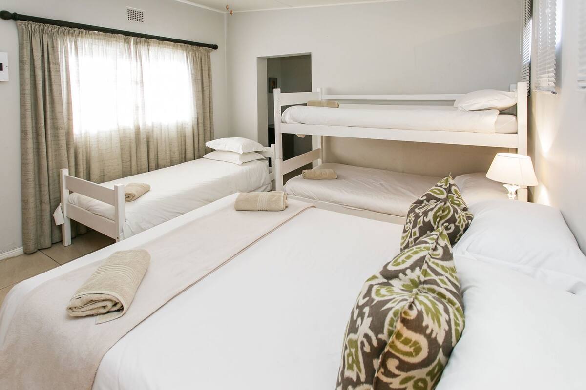 A hotel room with bunk beds, a single bed, a bathroom with a mirror, and a window with curtains.