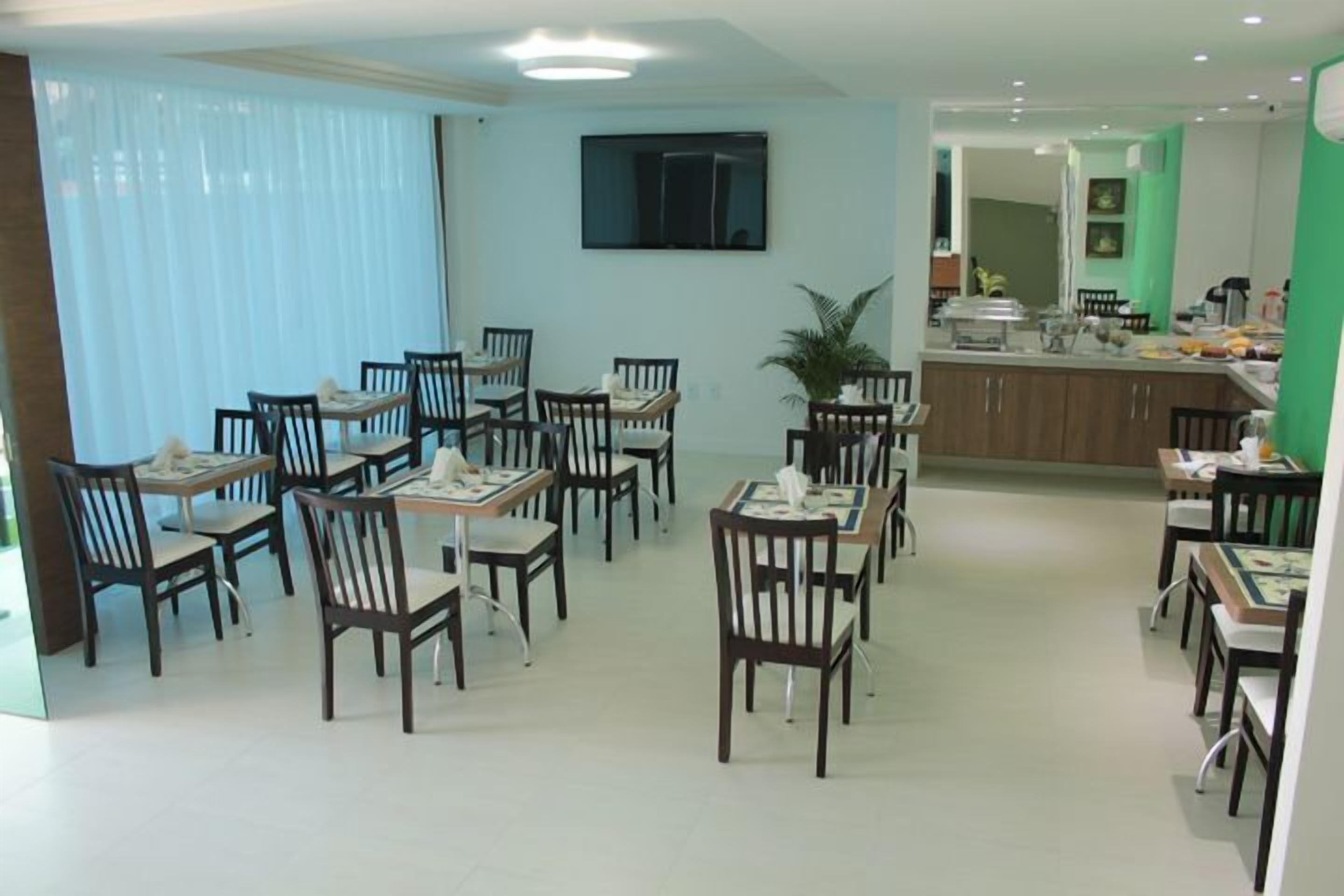 Uma área de refeições com várias mesas e cadeiras, uma televisão fixada na parede e uma cozinha ao fundo.