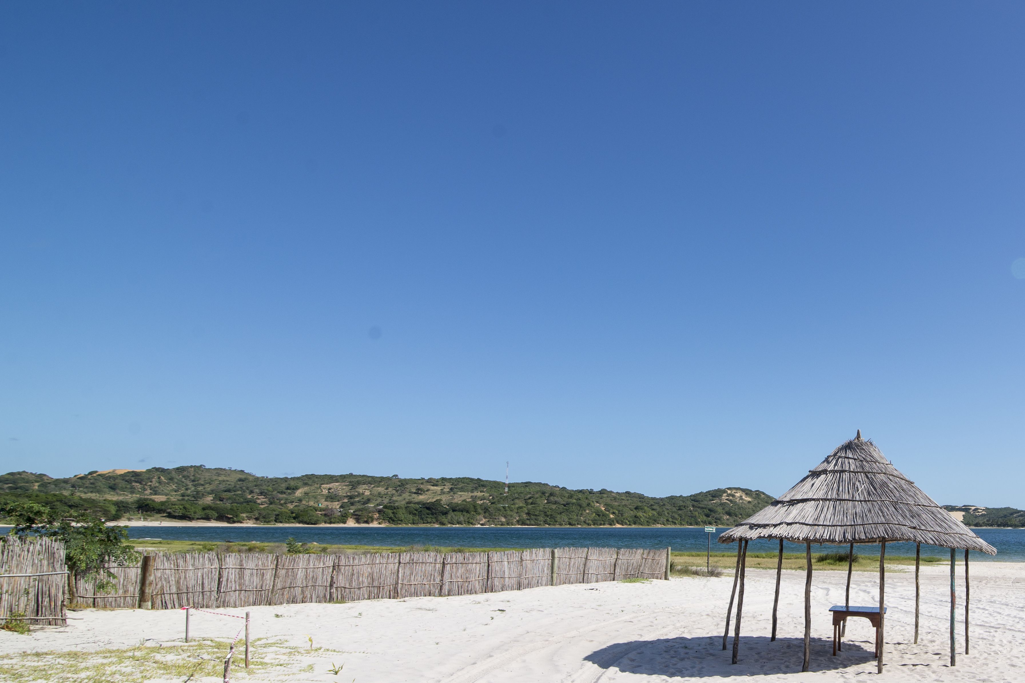 A beach with a thatched hut and a bench, surrounded by a wooden fence and a sandy area.