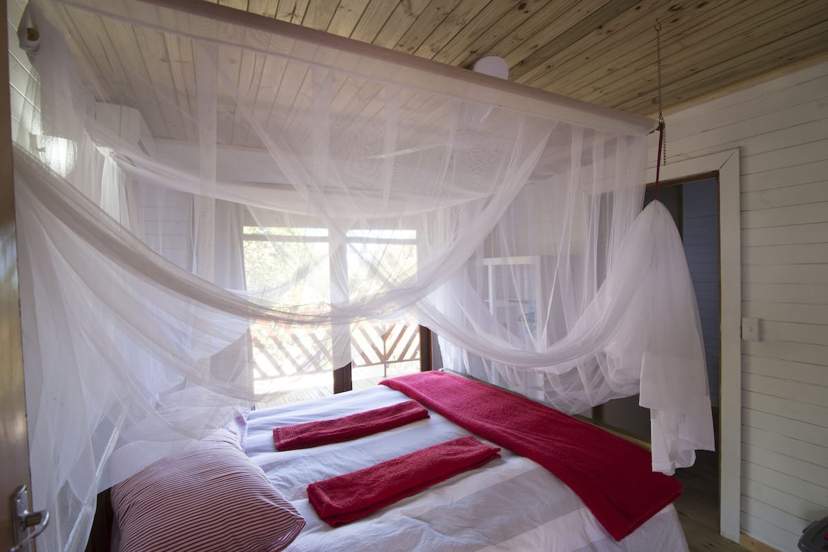 Een kamer met een bed, overdekt met een witte hemel, een raam met uitzicht, en een deur aan de rechterkant.
