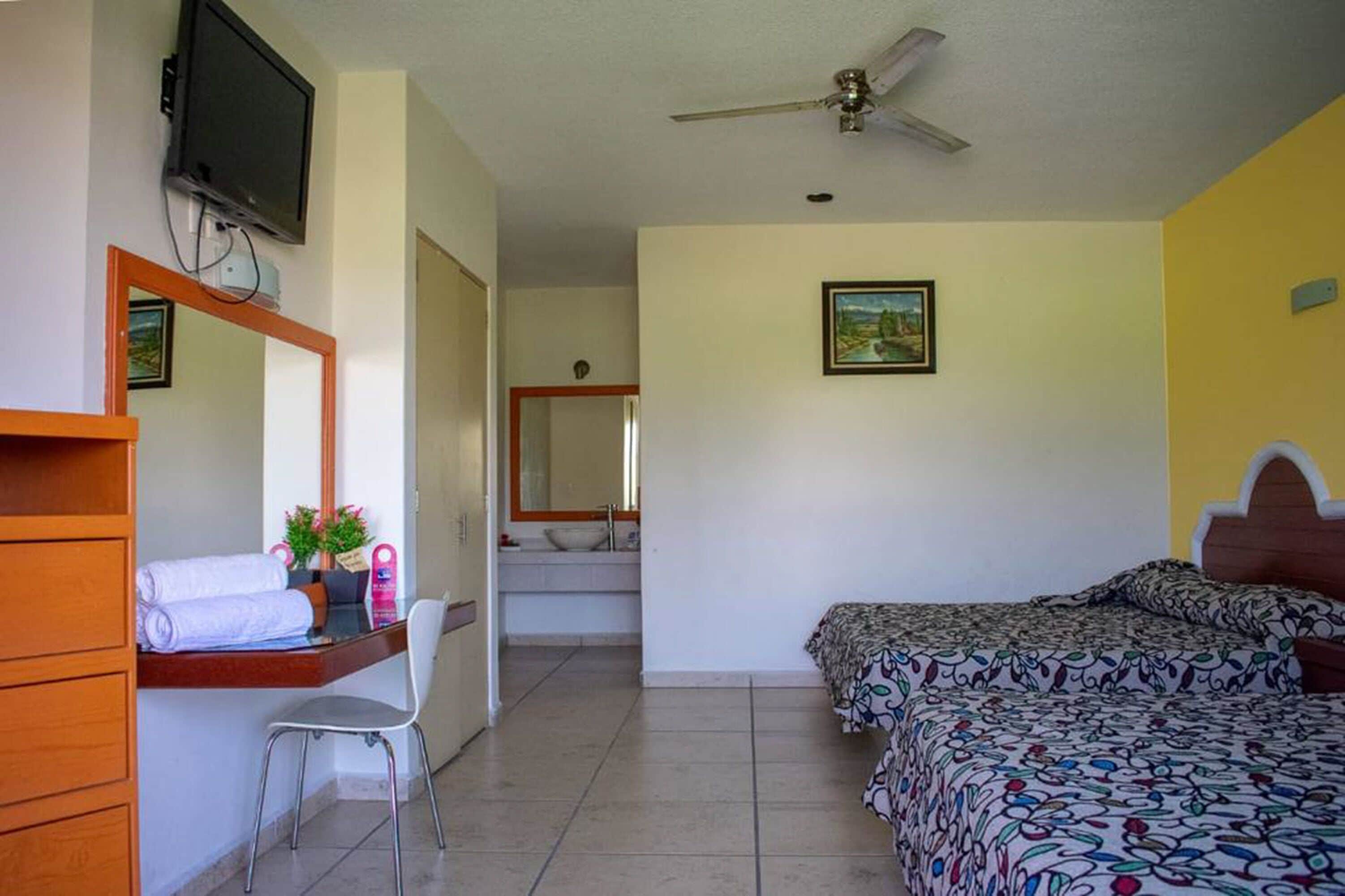 Un dormitorio con dos camas, un escritorio con silla, un televisor montado en la pared y un ventilador de techo.