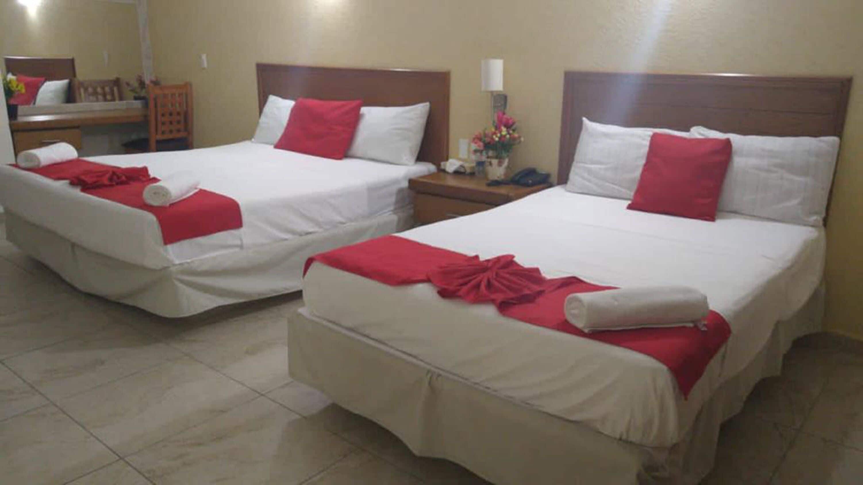 Two twin beds with white linens and red accents, a wooden headboard, and a small bedside table with a lamp and flowers.