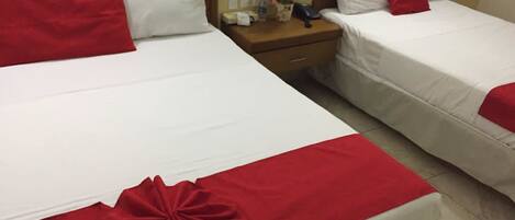 Two beds with white linens and red accents, a nightstand with a phone and a vase of flowers, and a wooden headboard.