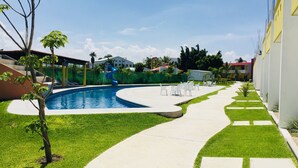 A swimming pool with a slide, surrounded by a grassy area and a paved walkway. - Hotel JS (Yautepec)