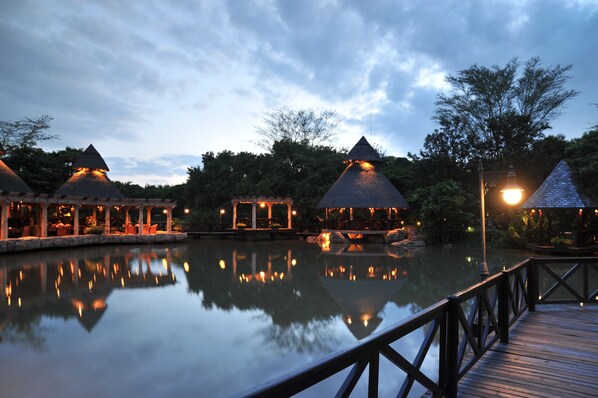 A tranquil lakeside resort with thatched-roof structures, wooden walkways, and a calm water body reflecting the buildings.