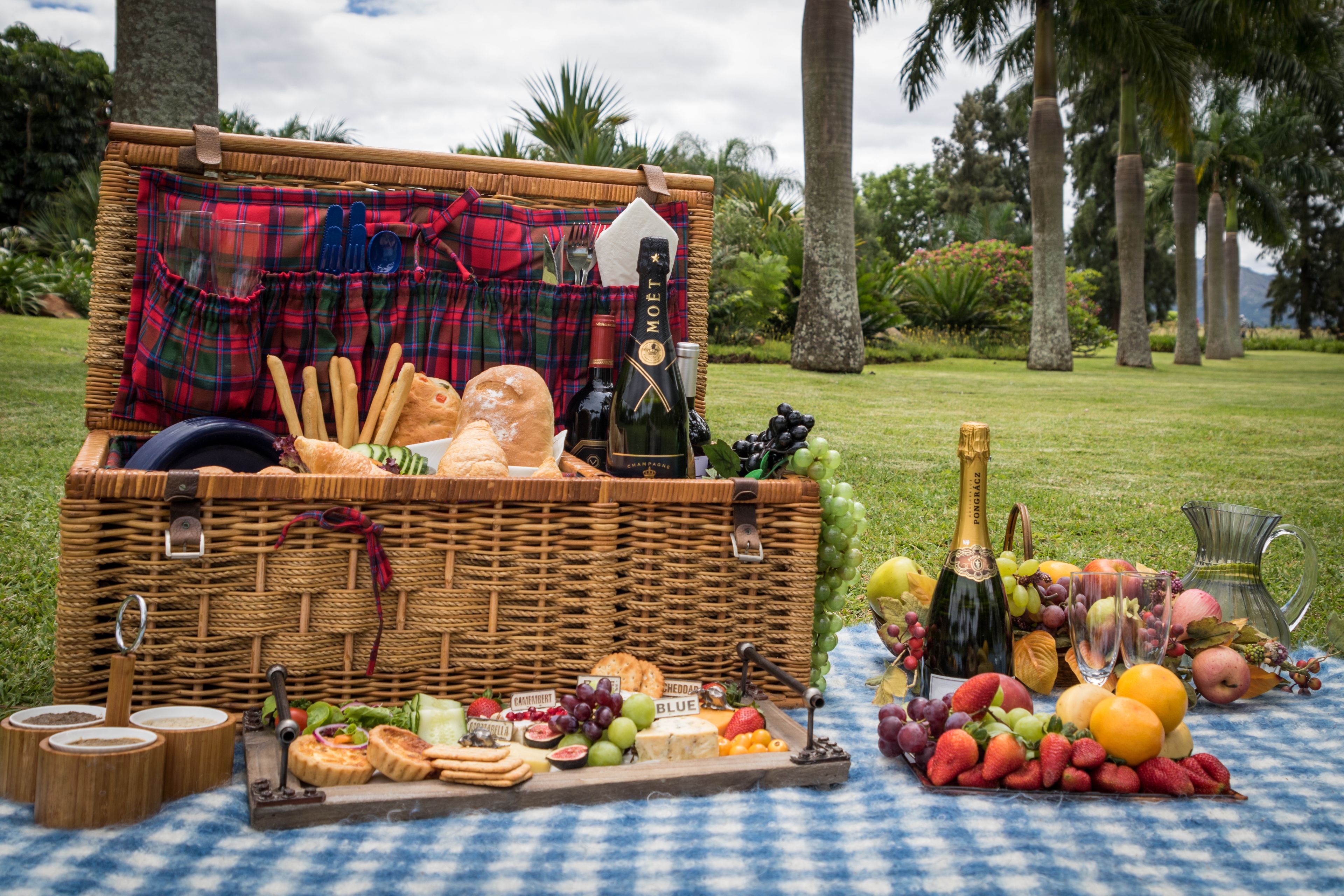 A picnic setup with a wicker basket containing bread, cheese, and a bottle of champagne, surrounded by fruits and snacks on a checkered tablecloth.