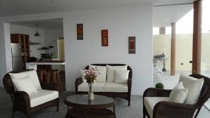 A modern living room with a sofa, two armchairs, and a coffee table. There is a kitchen in the background, and the room has large windows. - Casa de Playa Akas (Canoas de Punta Sal)