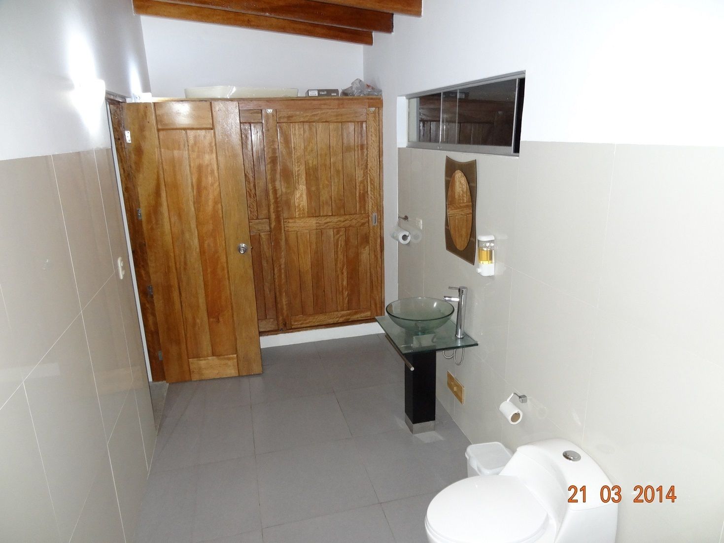 Un baño con puerta de madera, lavamanos de vidrio, inodoro y espejo.