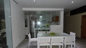 A modern dining area with a glass partition, white chairs, and a table with a fruit bowl. - Casa de Playa Akas (Canoas de Punta Sal)