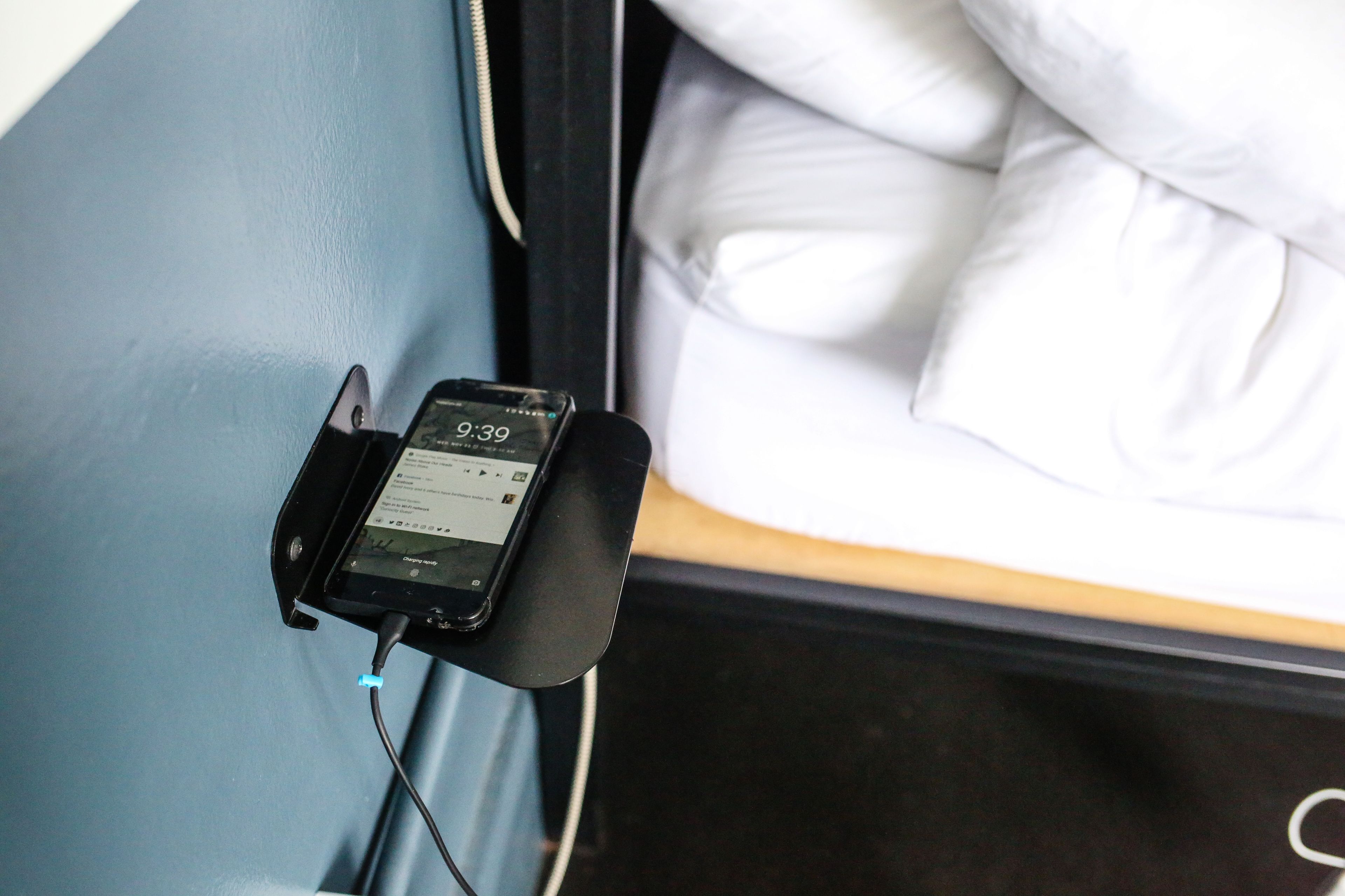 Un teléfono inteligente cargando sobre un soporte cerca de una cama con ropa de cama blanca.