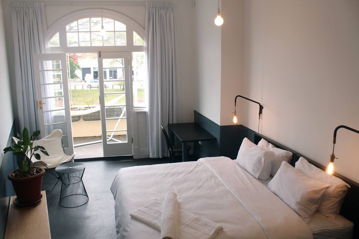 Habitación de hotel con cama, escritorio, silla, planta en maceta y vistas al exterior.