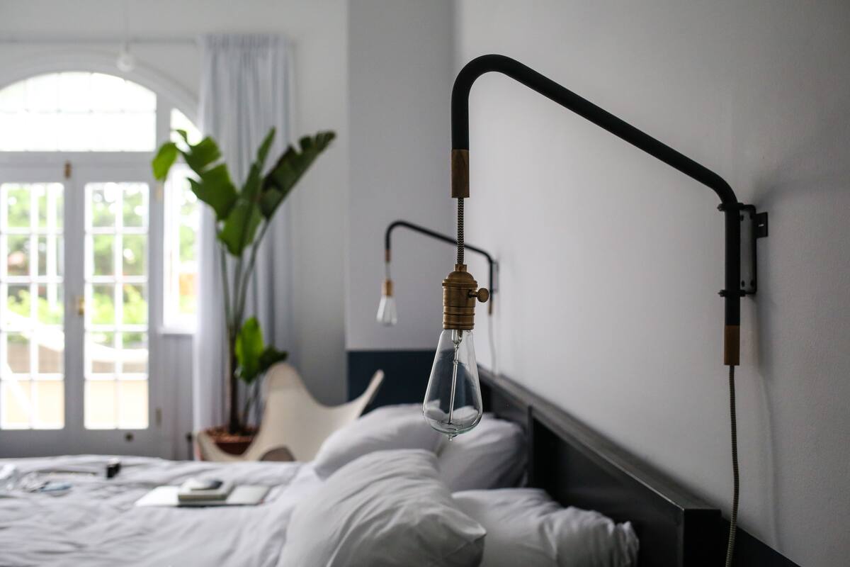 Un dormitorio con cama, lámpara colgante, planta en maceta y ventana con cortinas.