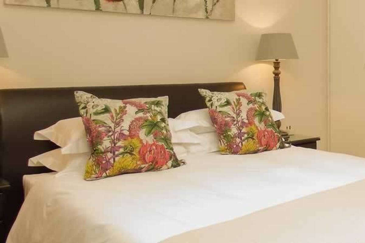 A neatly made bed with floral pillows, a bedside lamp, and a view into a bathroom with a sink and mirror.