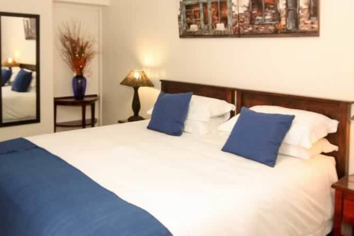 A neatly made bed with blue and white bedding, two bedside lamps, a nightstand, a mirror, and a framed picture on the wall.