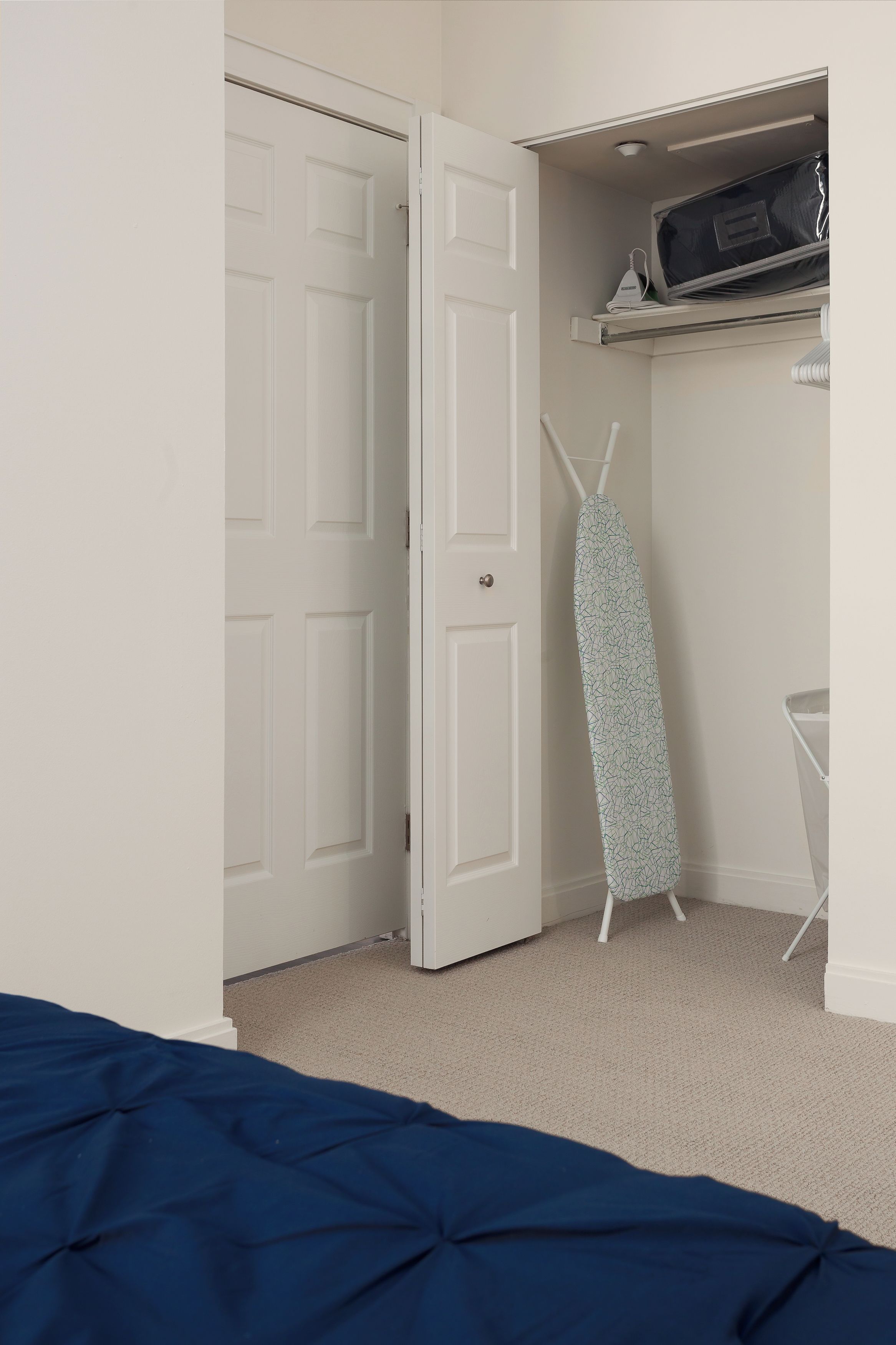 A bedroom with a bed, a door, an ironing board, and a shelf with a bag.