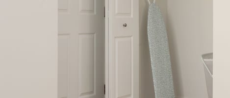 A bedroom with a bed, a door, an ironing board, and a shelf with a bag.