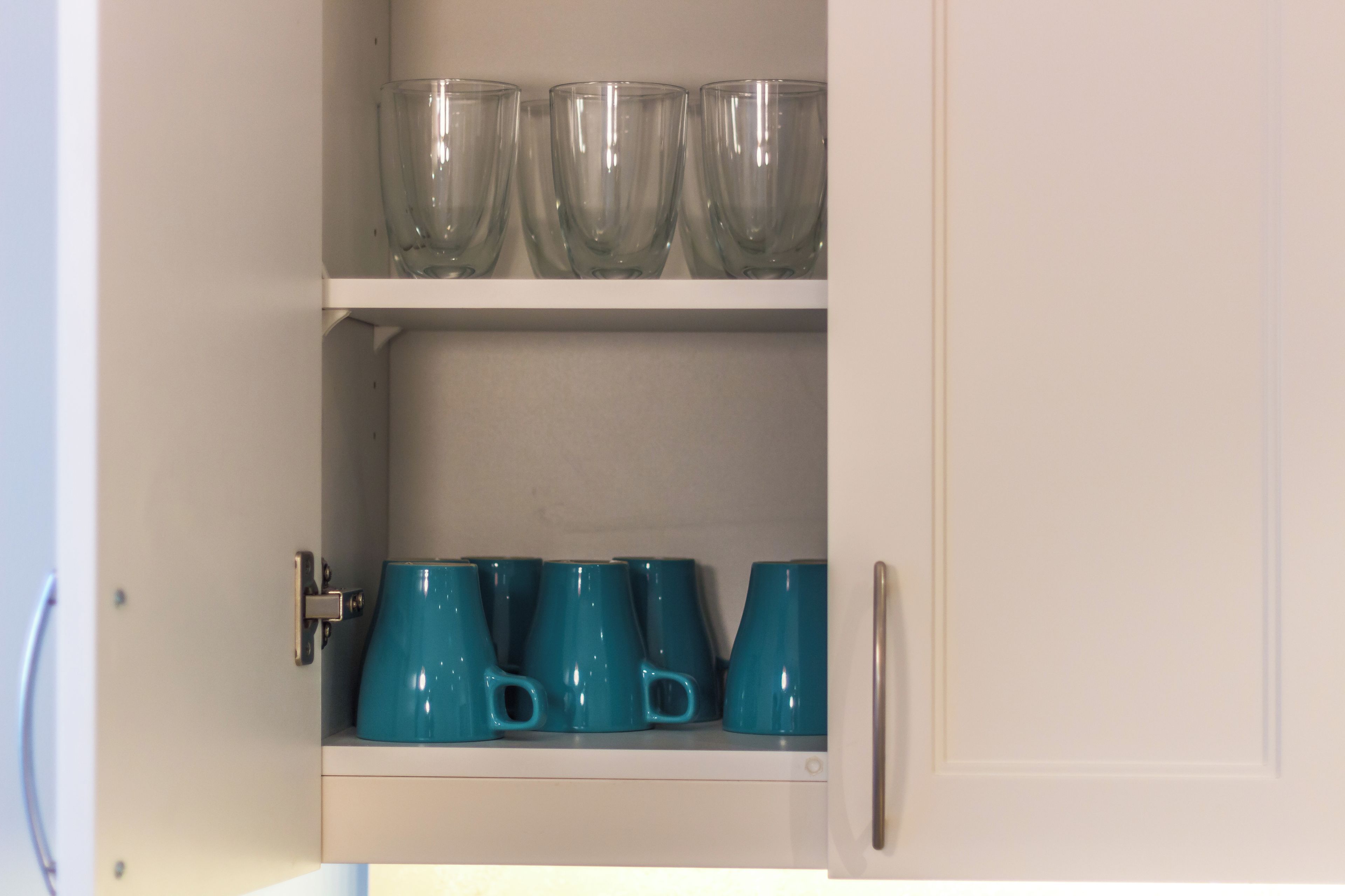 A cabinet with two shelves; the top shelf holds clear glass cups, and the bottom shelf has teal mugs.