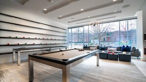 A modern indoor pool table area with a sofa and coffee table set, large windows, and a view of the outside. - Pelican Fully Furnished Apartments (Philadelphia)