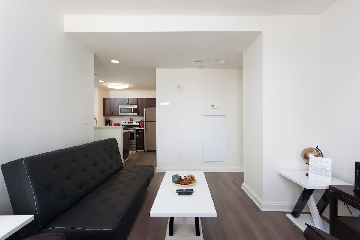 A modern living room with a black sofa, a white coffee table, and a kitchen in the background.