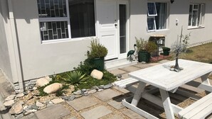 Un patio con una mesa blanca y bancos, rodeado por un camino de piedra y plantas en macetas.