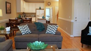 A living room with a gray sofa, two chairs, a coffee table with a green decorative bowl, a dining area with a table and chairs, and a kitchen with white cabinets and appliances. - Turtle Bay 10 3 Bedroom Holiday Home By My Ocean Rentals (Folly Beach)