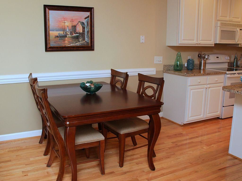 A dining room with a wooden table and chairs, a painting on the wall, and a kitchen area with white cabinets and appliances.