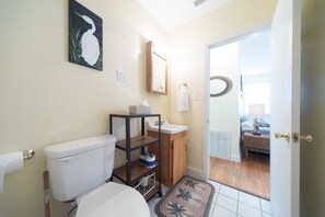 A bathroom with a toilet, sink, and shelves. There is a door leading to a bedroom.