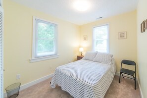 A bedroom with two windows, a bed with a polka dot bedspread, a nightstand with a lamp, and a chair.