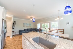 A modern kitchen with a granite countertop, stainless steel sink, and a built-in oven. There is a dining area with a bench and chairs, and a large window providing natural light. - E Ashley A 121 4 Bedroom Holiday Home By My Ocean Rentals (Folly Beach)