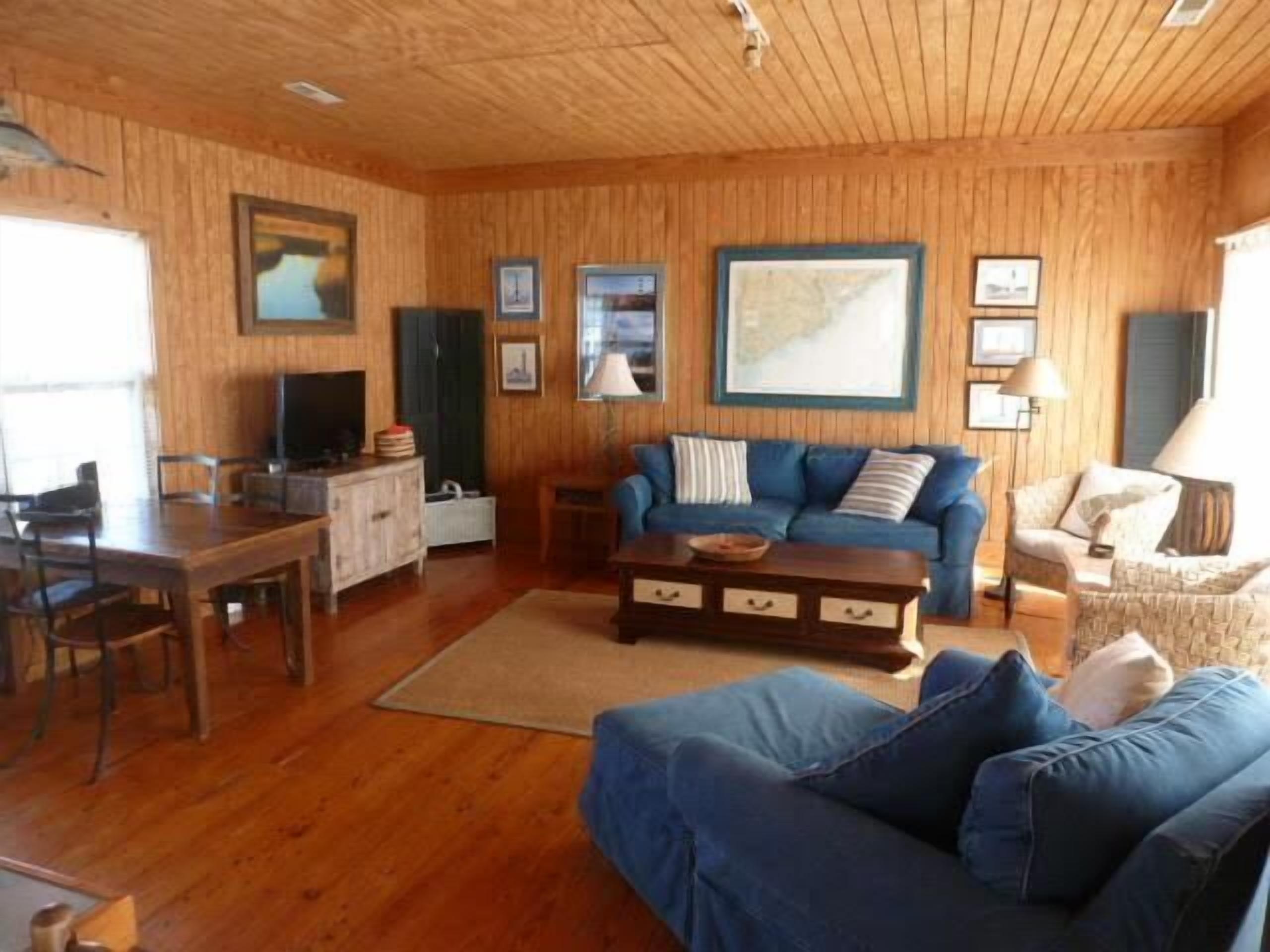 Un salon chaleureux aux murs et au plancher en bois, comprenant un canapé bleu, une table basse, une table à manger avec des chaises et un téléviseur.