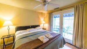 Camera d'albergo con un letto, una ventola a soffitto, due lampade da comodino e vista su un patio esterno con sedie.