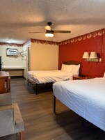 Habitación de hotel con dos camas, ventilador de techo, ventana pequeña con aire acondicionado, suelo de madera y pared roja con un borde floral.