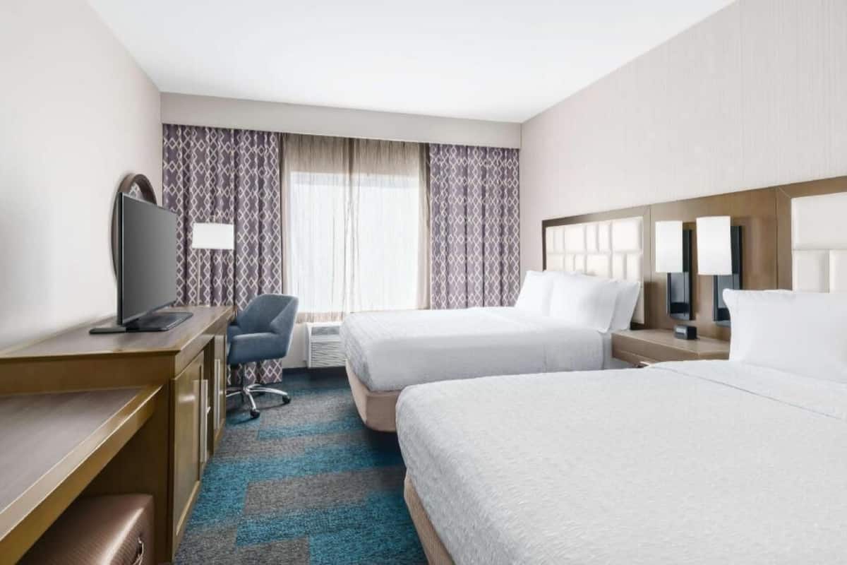 A hotel room with two beds, a desk, a chair, a TV, and a window with curtains.