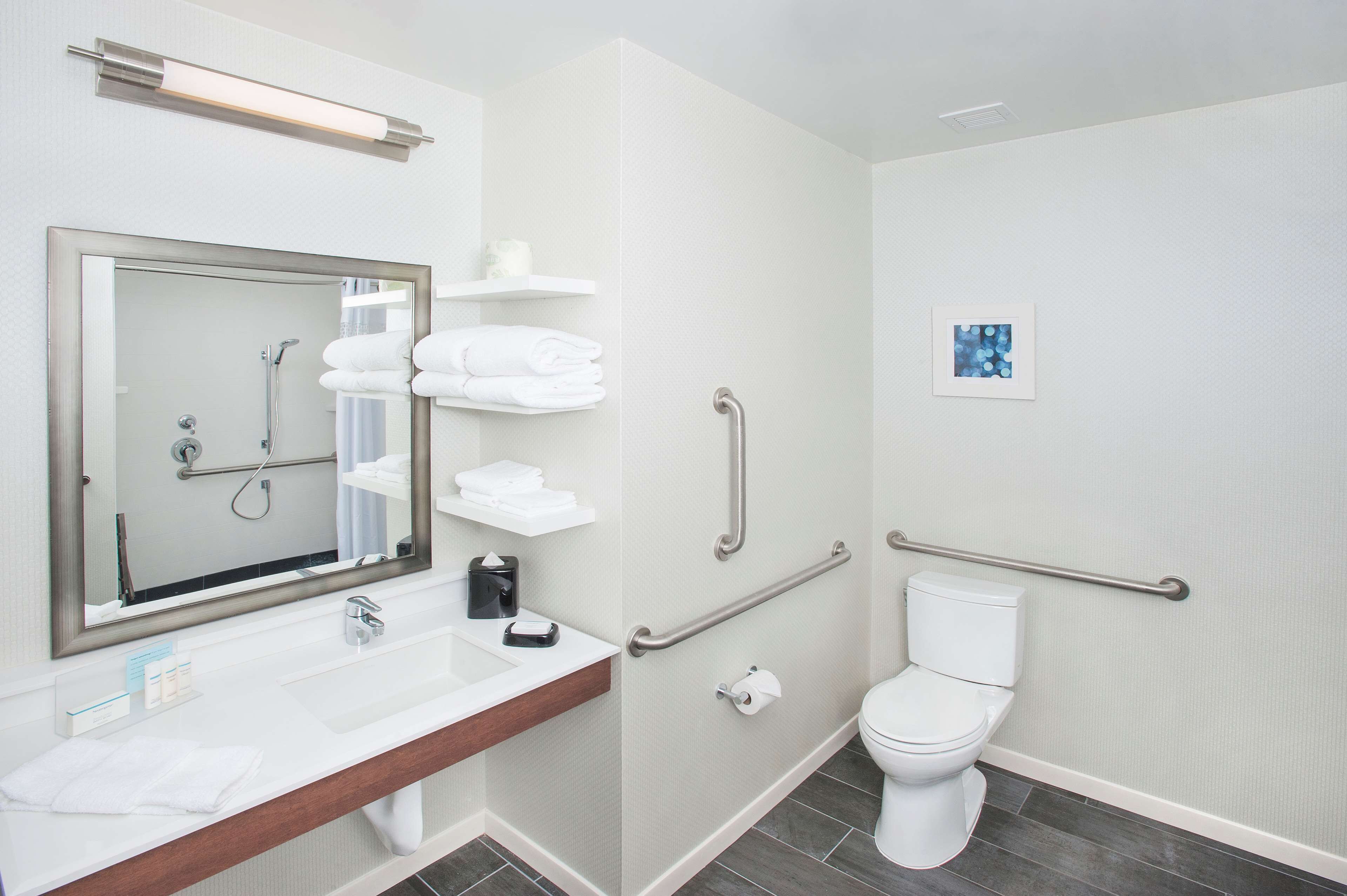 Ein modernes Badezimmer mit einem großen Spiegel, einem weißen Waschbecken und einer Toilette. Es gibt Handläufe und ein Regal mit Handtüchern.