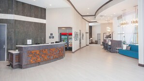 Un espace d’accueil moderne doté d’un plafond incurvé, d’un comptoir d’accueil avec un panneau décoratif en bois, d’un réfrigérateur rempli de boissons et d’un coin salon avec un canapé bleu et un lustre.