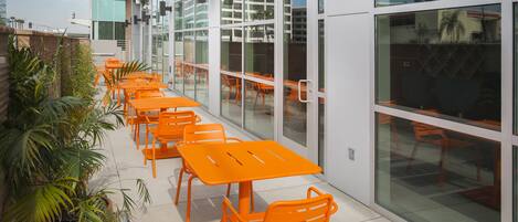 A modern outdoor seating area with orange tables and chairs, surrounded by glass walls and potted plants.