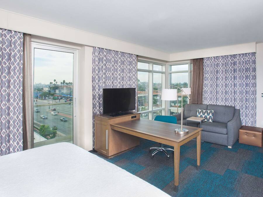 Habitación de hotel con una cama grande, un escritorio con televisor, un sofá y vistas a la ciudad.