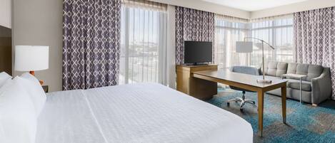 Habitación de hotel con una cama grande, un escritorio con televisor, un sofá y vistas a la ciudad.