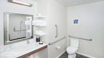 Un baño moderno con lavamanos, espejo y váter blancos. Incluye ducha con cabezal de ducha de mano y barras de apoyo para accesibilidad.