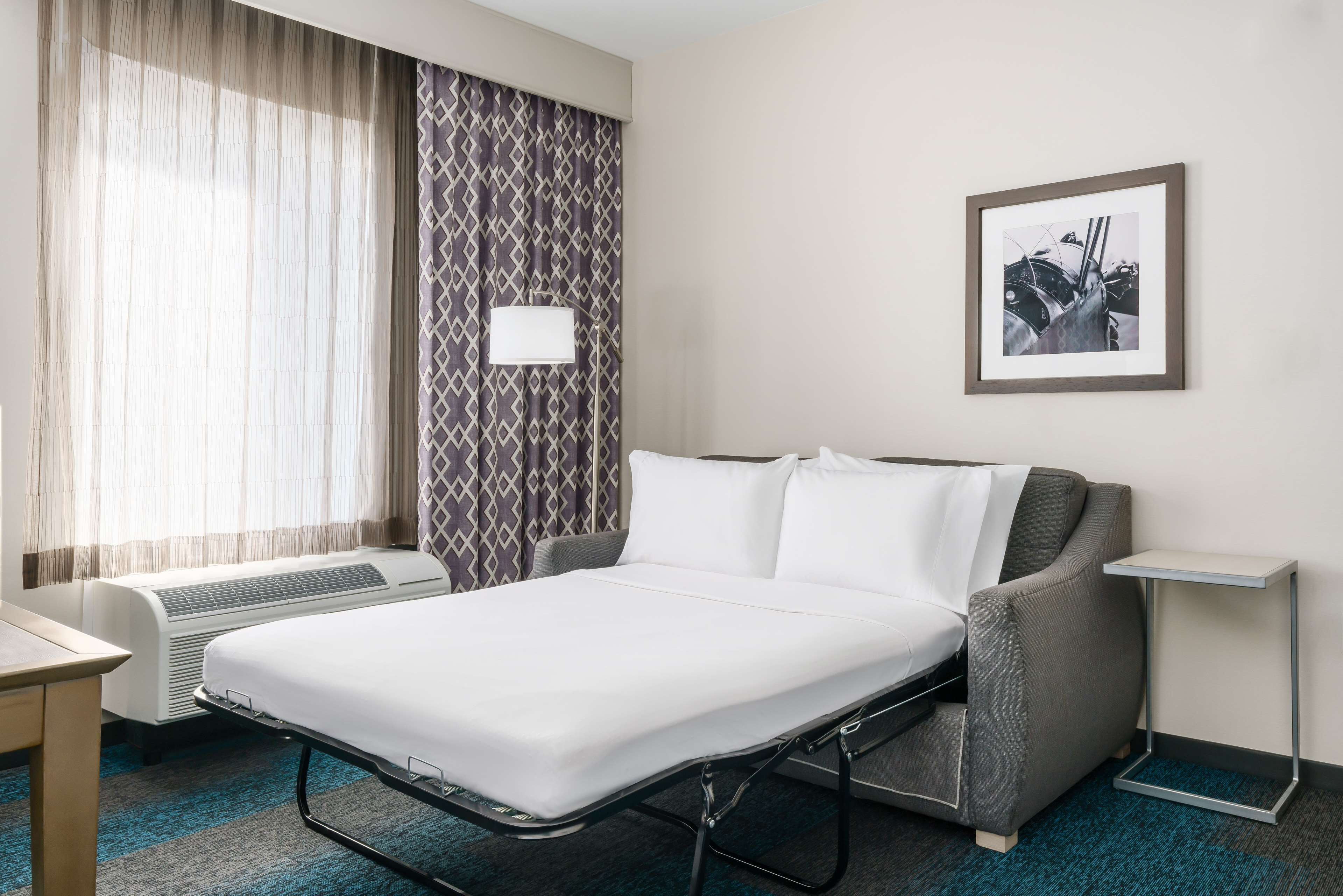 Une chambre d’hôtel avec un lit, un bureau, une chaise, une lampe fixée au mur et un tableau encadré accroché au mur.