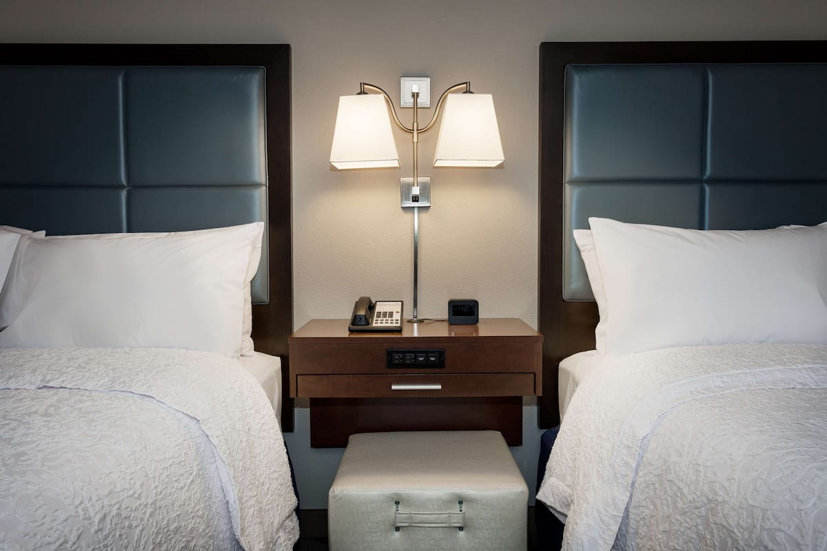 A hotel room with two beds, a nightstand with a lamp, a phone, and a digital clock.