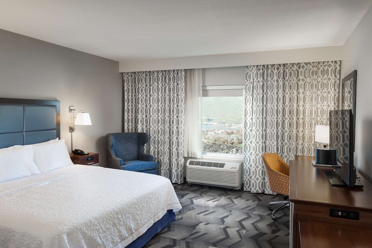 A hotel room with a large bed, a desk with a TV, a chair, a window with curtains, and an air conditioning unit.