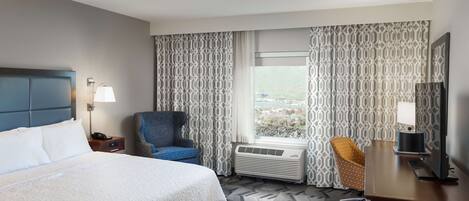 Ein Hotelzimmer mit einem großen Bett, einem Schreibtisch mit Fernseher, einem Sessel, einem Fenster mit Vorhängen und einer Klimaanlage.