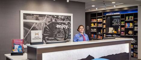 A hotel reception desk with a large framed picture of a bicycle, a sign reading "Hilton HH Honors," and a display of snacks.