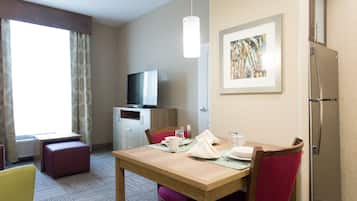 A hotel room with a wooden dining table, red chairs, a television, a refrigerator, and a framed artwork on the wall.