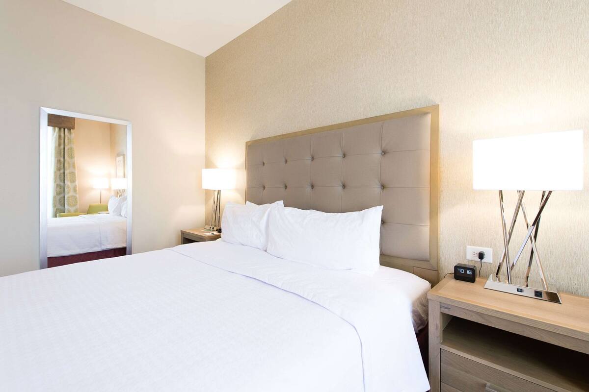 Una habitación de hotel con una cama grande, una mesita de noche con lámpara, una mesita con un reloj y un espejo en la pared.