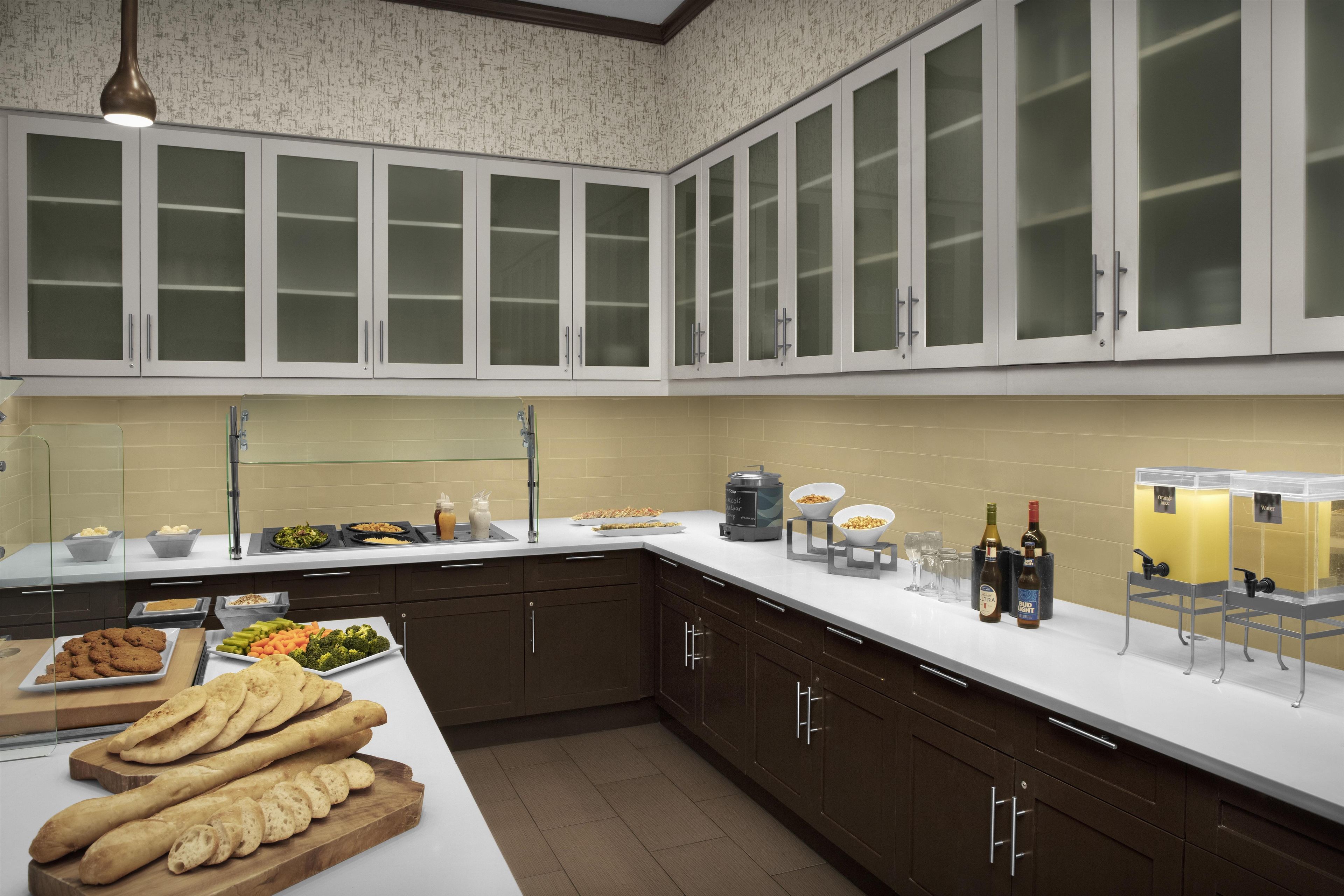A modern kitchen with dark brown cabinets, a white countertop, and a variety of food items displayed.