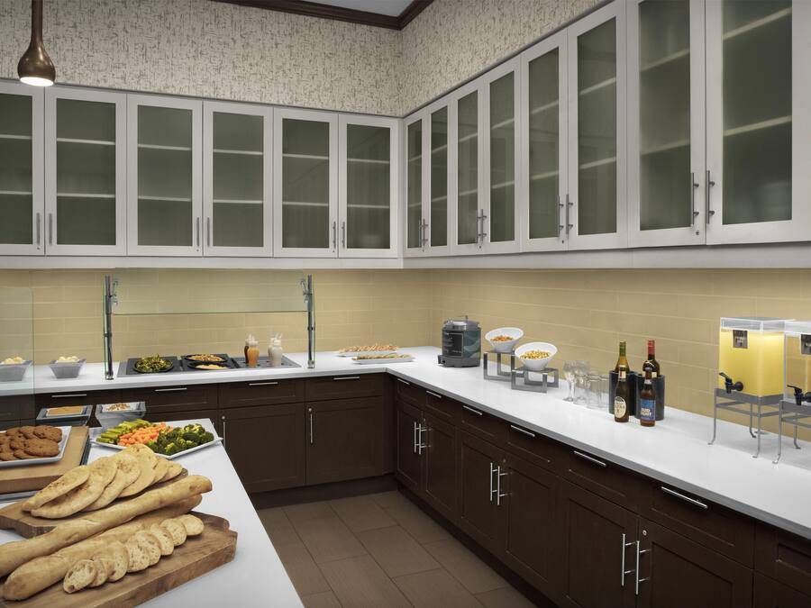A modern kitchen with dark brown cabinets, a white countertop, and a variety of food items displayed.